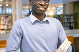 Optimistic African American man in eyeglasses with stack of books in hands standing in university library