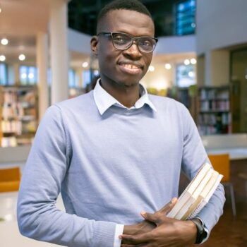 Optimistic African American man in eyeglasses with stack of books in hands standing in university library